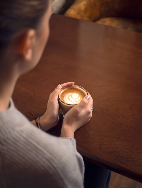 Kaffeelatte in einem hohen Glas. Kaffee auf Holztisch. Steinmauer. Vintage-Interieur. Frau hält Tasse Kaffee, junges Mädchen trinkt Kaffee. Tasse auf Holztisch vorhanden. Retro-Foto.  - Foto, Bild