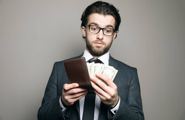 muž v obleku a brýlích vytáhne peníze z peněženky na šedém pozadí - Fotografie, Obrázek