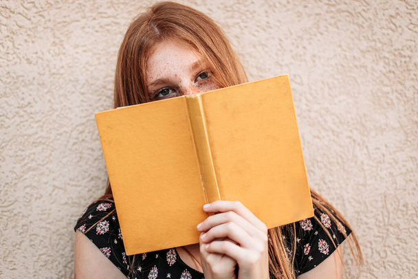 Teen ginger student girl holding a book, face hidden with a book - 写真・画像