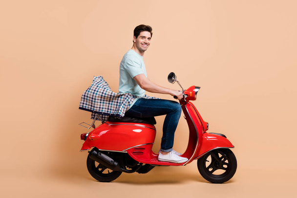 Profile side view portrait of attractive cheerful guy driving red moped spending time way road tour isolated over beige pastel color background - Фото, изображение