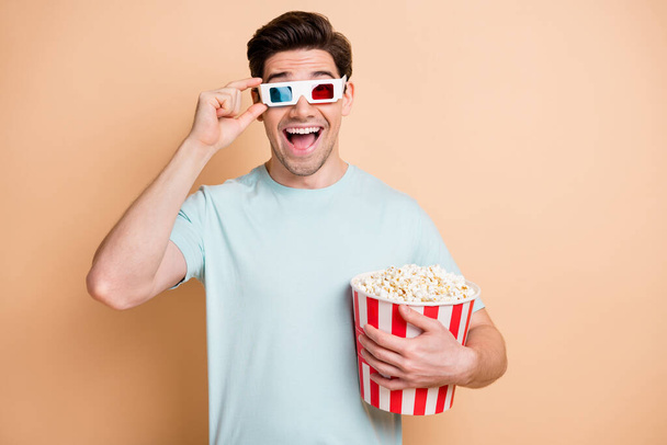 Portrait of nice cheerful amazed guy eating pop corn watching film touching 3d specs isolated over beige pastel color background - Fotoğraf, Görsel