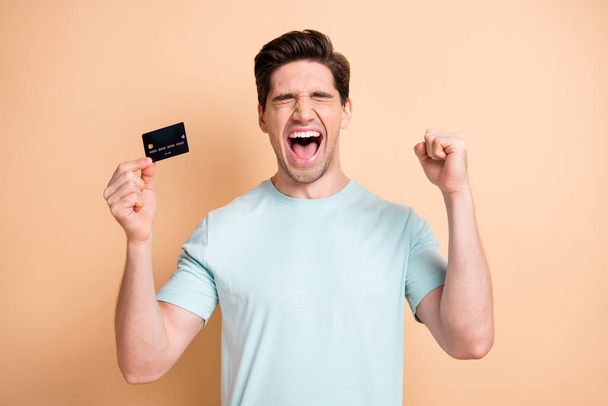 Portrait of nice lucky cheerful guy holding in hand bank card celebrating success having fun isolated over beige pastel color background - Foto, immagini