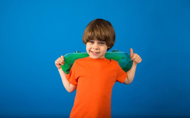 πορτρέτο ενός χαρούμενου μικρού αγοριού σε ένα πορτοκαλί T-shirt κρατώντας ένα skateboard σε μπλε φόντο με ένα αντίγραφο του χώρου - Φωτογραφία, εικόνα