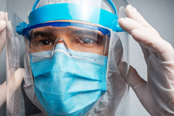 Close-up portrait of young doctor wearing PPE suit against coronavirus and covid-19, on background of grey wall. - Photo, Image