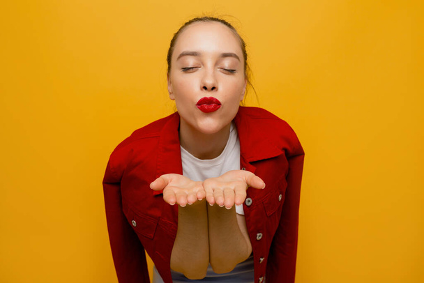 Wonderful happy woman with red lips sending a kiss with closed eyes over yellow backdrop. High quality photo - Фото, изображение