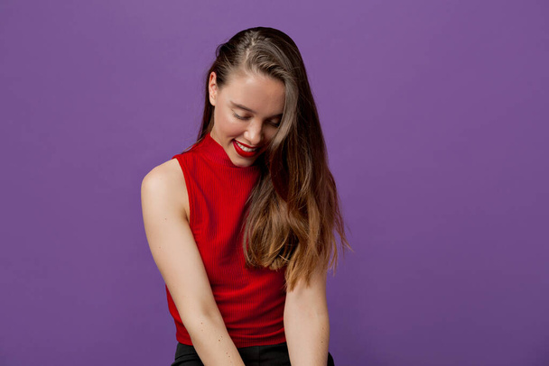 Studio portrait of attractive pretty woman with brown hair wearing red top looking down and smiling over violet backdrop. High quality photo - Photo, Image