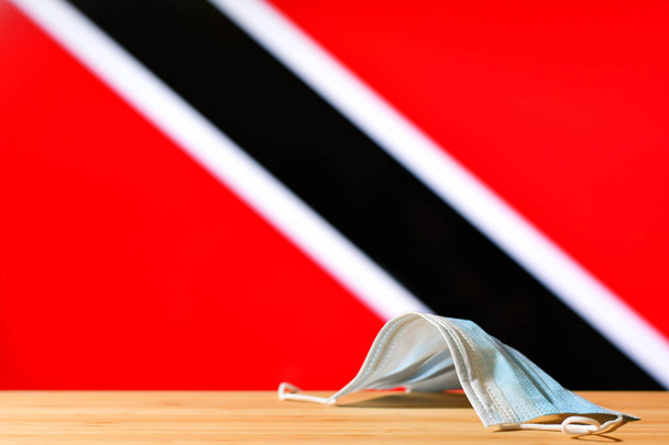 Una maschera medica si trova sul tavolo sullo sfondo della bandiera di Trinidad e Tobago. Il concetto di un regime di maschera obbligatoria per i residenti del paese e turisti a Trinidad e Tobago durante una pandemia. - Foto, immagini
