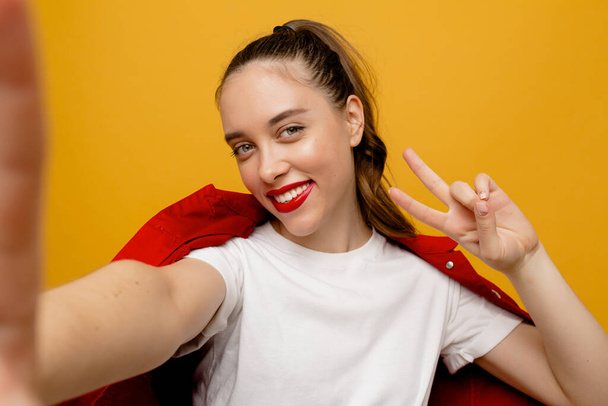 Χαμογελαστή ευτυχισμένη κοπέλα με κόκκινα χείλη φορώντας κόκκινο σακάκι κάνοντας selfie με πινακίδα ειρήνης σε απομονωμένο φόντο. Υψηλής ποιότητας φωτογραφία - Φωτογραφία, εικόνα