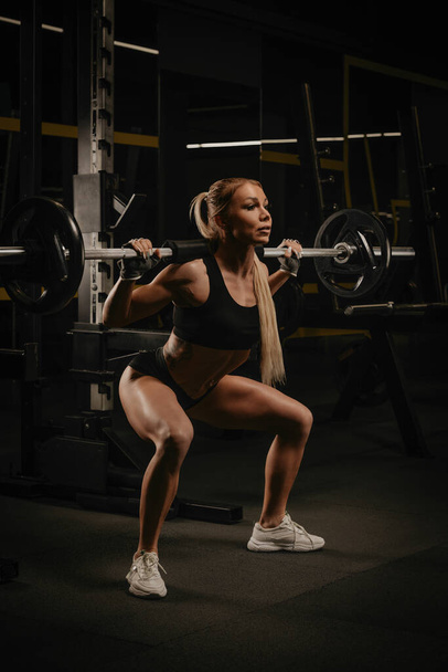 A fit woman with blonde hair is squatting with a barbell near the squat rack in a gym. A girl is doing a leg workout. - Foto, immagini