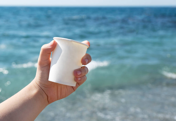 Weibliche Hand hält weiße Kaffeetasse aus Karton auf blauem Meeresgrund, Kaffee zum Mitnehmen draußen, aktives Leben und Freizeitkonzept - Foto, Bild
