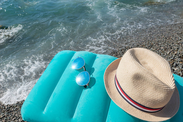 Almohada de natación inflable azul, gafas de sol brillantes y sombrero de sol se encuentran en la playa cerca del mar con olas. Concepto de viaje, planificación vacaciones de verano en el mar. - Foto, imagen