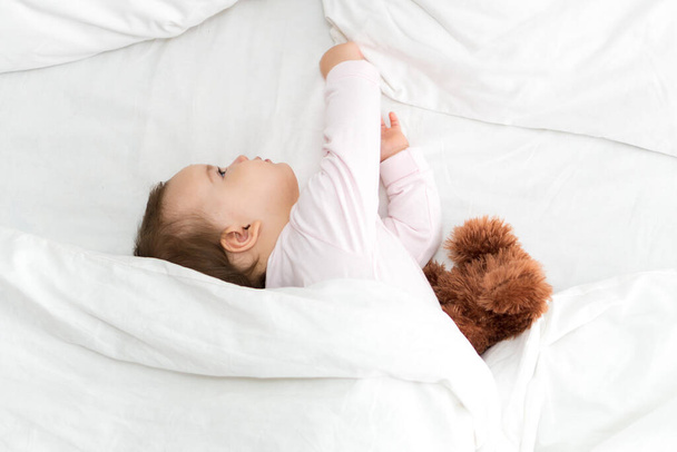 Authentic portrait cute caucasian little infant chubby baby girl or boy in pink sleepy upon waking with teddy bear looking at camera in white bed. Child care, Childhood, Parenthood, lifestyle concept - Foto, Imagem