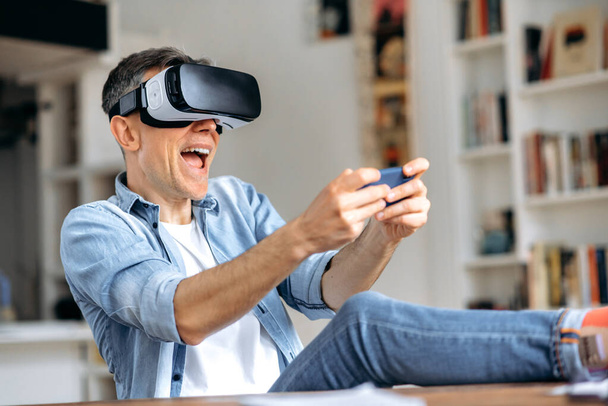 Middle aged caucasian man, freelancer, wearing virtual reality goggles during working break. Excited man testing VR glasses while sits at the desk, smartphone using with VR headset, virtual reality - Foto, afbeelding