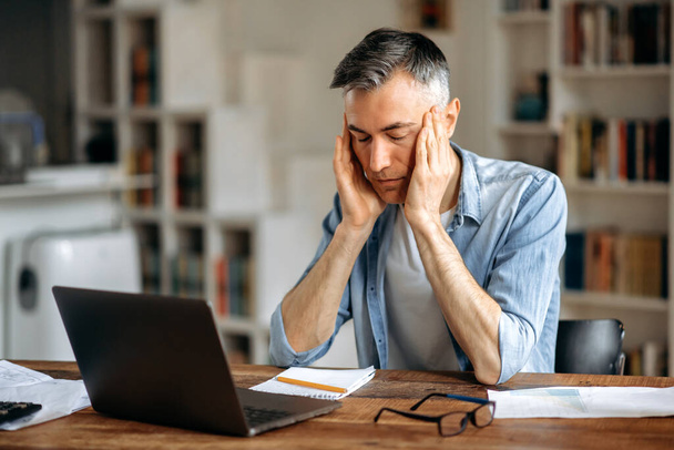 Mature caucasian gray-haired exhausted man, manager or freelancer, sitting at work desk, holding his head with his hands, tired of working in laptop, have a headache, migraine, need rest - Foto, Bild