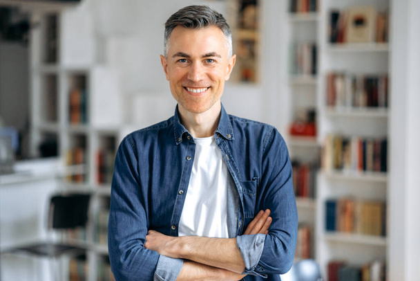Portrait of an attractive mature gray haired caucasian man, freelancer or manager, standing near his work desk in denim shirt with arms crossed, looking and smiling friendly at camera - Photo, Image