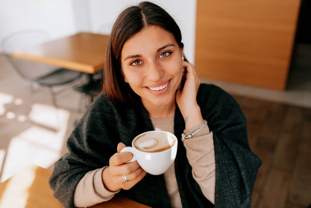Γοητευτική λατίνα μελαχρινή γυναίκα με μακριά μαλλιά χαμογελώντας πίνοντας πρωινό καφέ στο φως του ήλιου. Υψηλής ποιότητας φωτογραφία - Φωτογραφία, εικόνα