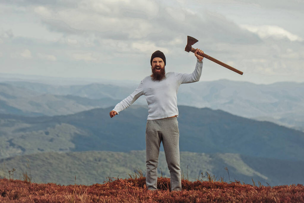 Handsome bearded man with axe. Brutal lumberjack hipster with beard and moustache stands with axe on mountain top. - Foto, immagini