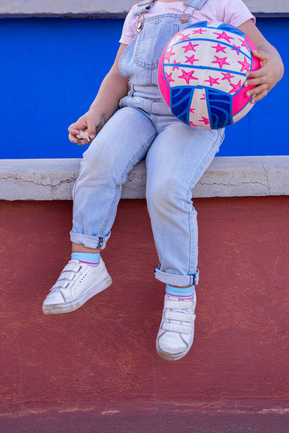 Legs of an unrecognizable two-year-old girl in denim overalls, sitting on the bleachers, holding a ball. - Photo, Image