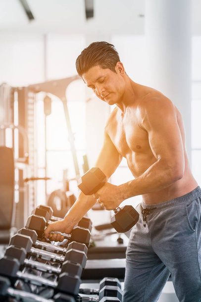 gym and fitness background with light flare of close up dumbbell with hand of athletic bodybuilder man lifting from dumbbell rack in gym and futness club - Fotografie, Obrázek
