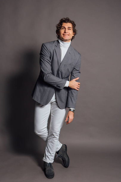 Fashion portrait of a tall handsome man dressed in white turtleneck, white jeans and grey jacket posing on the grey background - Foto, afbeelding