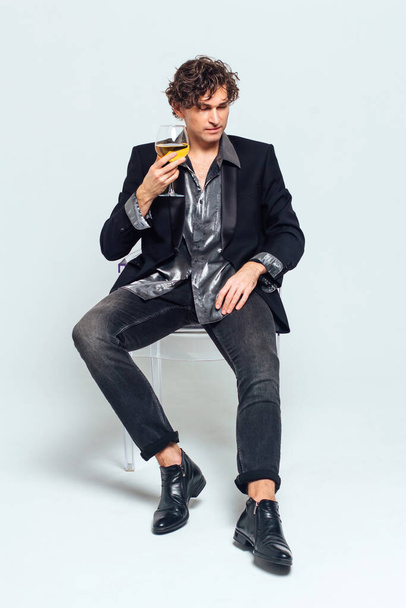 Tall handsome confident young man in a jacket sitting on the chair and holding glass of wine over white background - Foto, Imagem