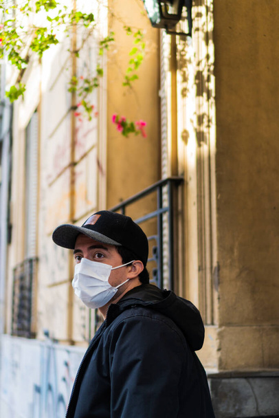 латиноамериканец смотрит на горизонт, идя по улицам Сан-Тельмо Буэнос-Айреса в хирургической маске, заботясь о вирусе Ковида. черный колпак и куртка, перед желтым домом - Фото, изображение