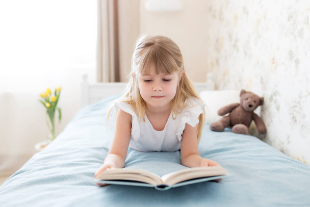 Ein kleines Mädchen legt sich auf das Bett im stilvollen Schlafzimmer, liest ein blaues Buch und macht Hausaufgaben. Bildung, häusliches Schulkonzept. Gelbe Tulpen in der Vase neben dem Bett - Foto, Bild