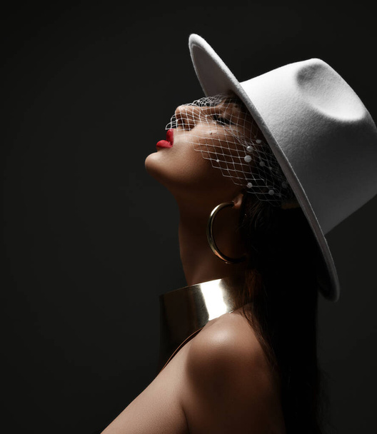 Profile of rich woman in white hat with veiling and massive metal collar and earrings holding head up with eyes closed - Fotó, kép