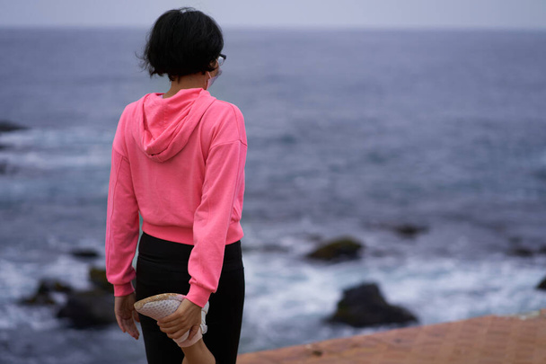 Mujer con máscara realiza estiramientos de piernas antes de hacer ejercicio mientras mira al mar. Nueva normalidad. sesión deportiva - Foto, Imagen