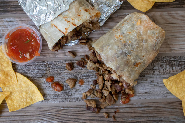Boven een carne asada beef burrito doormidden gesneden met vlees eruit. Het staat op een houten tafel met chips en salsa. - Foto, afbeelding