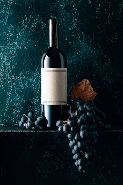 Bottle of red wine with the old empty label. Red wine and blue grapes with dried up vine leaves on an old dark blue background. Copy space. - Photo, Image