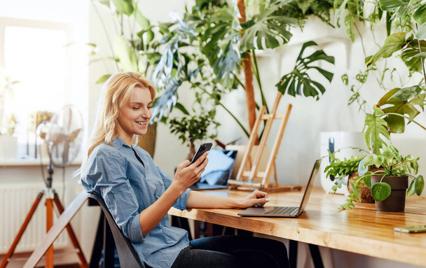 Молодая офисная работница со смартфоном и ноутбуком в офисе - Фото, изображение