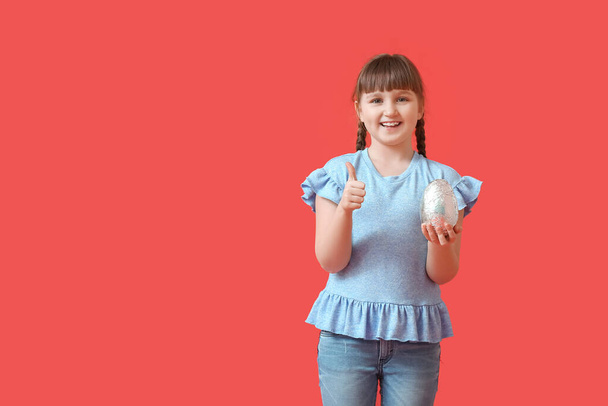 Милая маленькая девочка с шоколадным пасхальным яйцом показывает большой палец вверх на цветном фоне - Фото, изображение