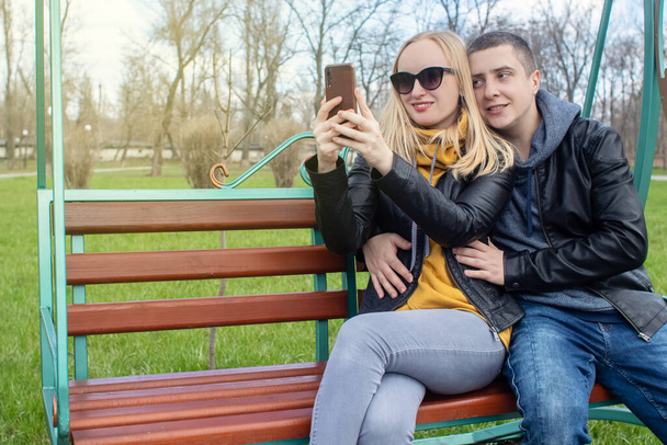 Οι σύζυγοι βγάζουν σέλφι ενώ κάθονται σε μια κούνια στο πάρκο. Ευτυχισμένο ζευγάρι που βγάζουν φωτογραφίες τον εαυτό τους. Η έννοια της ευτυχισμένης οικογένειας, ποζάρει και αιχμαλωτίζει τη στιγμή. - Φωτογραφία, εικόνα