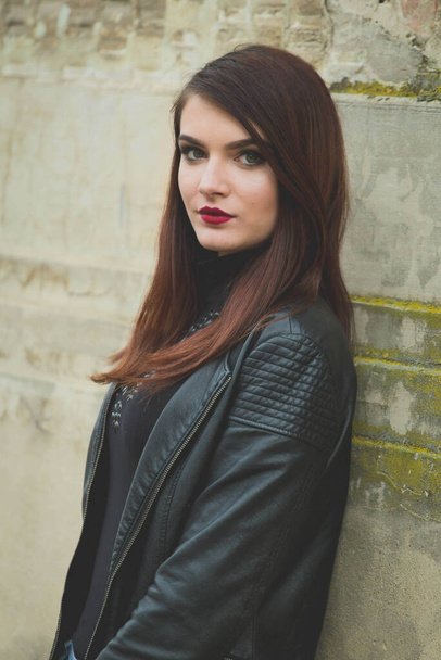 A vertical shot of a young brunette female in a leather coat posing in front of an old building - Foto, immagini
