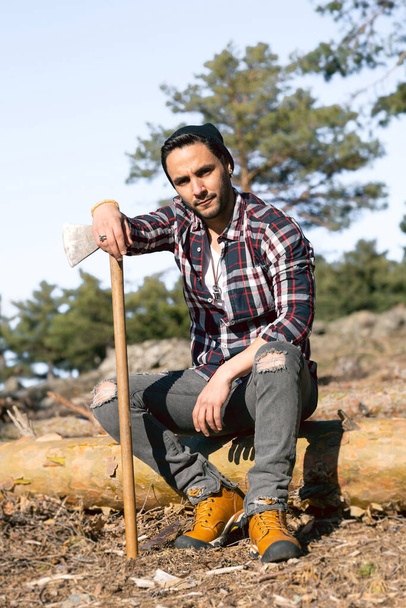 A closeup of a Spanish lumberman sitting on a wooden log and leaning on his ax - Valokuva, kuva