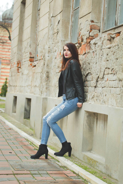 A vertical shot of a young brunette female in a leather coat posing in front of an old building - Photo, image
