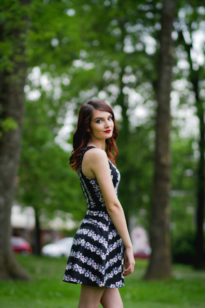 A vertical shot of a young brunette female in a floral dress posing in a park - Zdjęcie, obraz