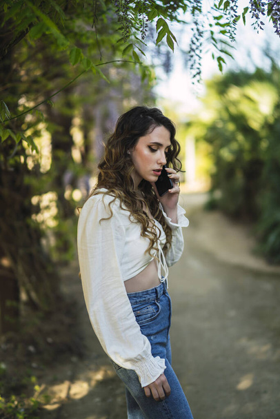 A vertical shot of a beautiful Spanish Caucasian woman with curly hair walking in a park, talking on her smartphone - Foto, Imagen