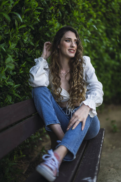 A beautiful Caucasian woman from Spain in blue jeans posing on a bench next to a hedge - Valokuva, kuva