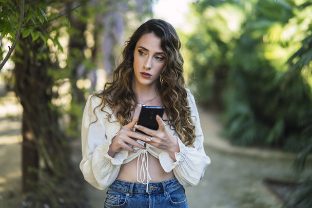 A shallow focus shot of a beautiful Spanish Caucasian woman with curly hair walking in a park with her smartphone - Фото, изображение