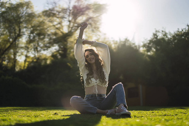 A beautiful Caucasian woman from Spain in white top and jeans sitting on grass and stretching on grass - Φωτογραφία, εικόνα