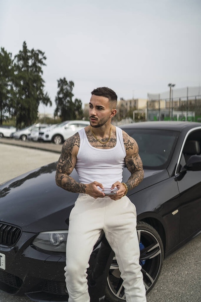 A vertical shot of a sexy tattooed male using his phone leaning on his car - Zdjęcie, obraz