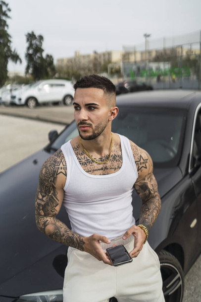 A vertical shot of a sexy tattooed male holding his phone leaning on his car - 写真・画像