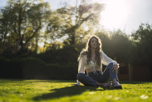A beautiful Caucasian woman from Spain in white top and jeans sitting on grass in a sunny park - Foto, Bild