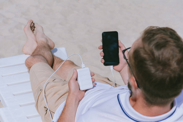 A man lies on a sun lounger with a mock-up of a smartphone and is charging from a Power Bank. Against the background of sand - Photo, image