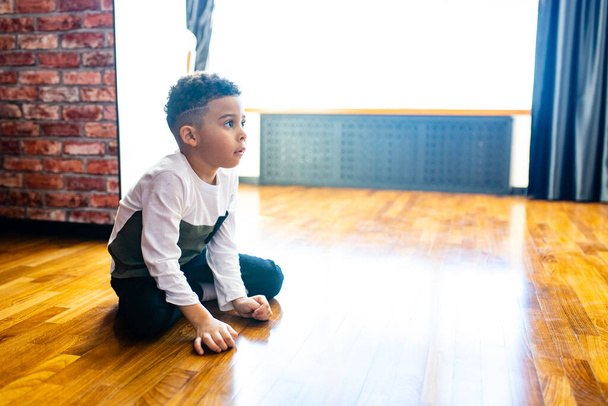 ethnic hispanic curly son sitting on floor warm brown clean safety parquet at home - Foto, Imagen