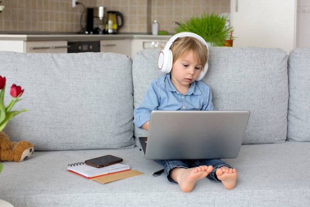 Sweet blond child, boy in casual clothes, woring on computed at home, with headphones and phone - Foto, Imagen