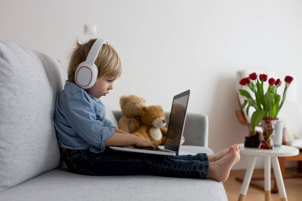 Sweet blond child, boy in casual clothes, woring on computed at home, with headphones and phone - Φωτογραφία, εικόνα