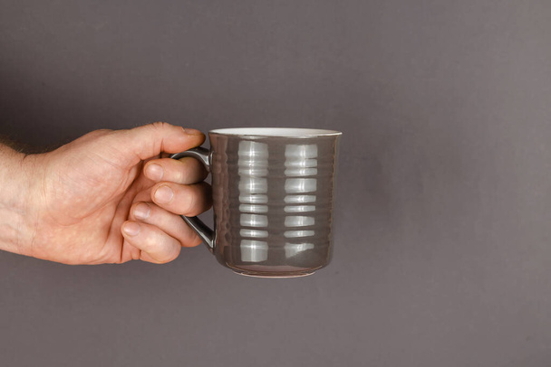 A hand and a mug on a gray background. A man is holding a gray ceramic mug. Side view. - Фото, изображение